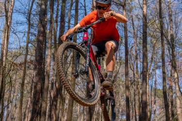 A mountain biker in an orange shirt and helmet performs a jump over a dirt ramp in a wooded area, surrounded by tall trees and clear blue skies. Lakeshore Bike Park mountain bike trail.