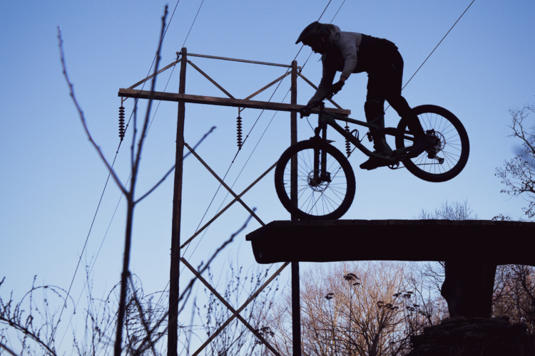 A silhouette of a mountain biker performing a jump over a wooden ramp, with power lines in the background and a clear blue sky above. Surrounding vegetation is partially visible in the foreground.