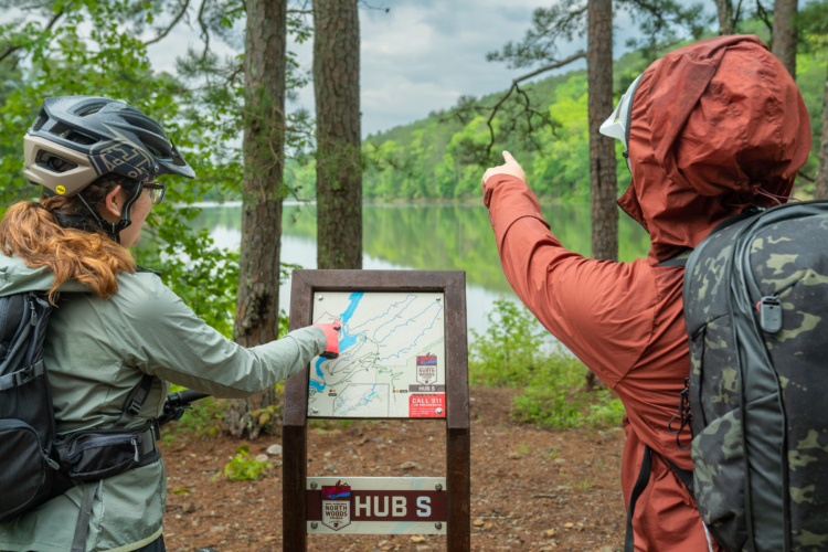 Two individuals, one wearing a helmet and the other dressed in a rain jacket, are studying a hiking map at a lakeside location. The person on the left points at the map while the other gestures toward the landscape in front of them. Lush greenery surrounds the area, and the calm lake reflects the trees and cloudy sky. A sign displaying "HUB S" is visible next to the map.