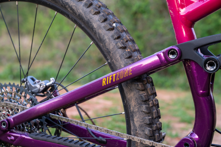 Close-up view of a purple mountain bike frame with the text "RIFTZONE" visible, alongside a dirty rear tire and components, set against a blurred green outdoor background.