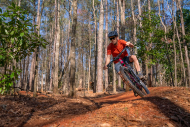 A mountain biker in an orange shirt and black helmet skillfully navigates a dirt trail surrounded by tall trees in a lush forest. The rider is mid-motion, leaning into a turn on the bike, with sunlight filtering through the leaves above. Lakeshore Bike Park mountain bike trail.