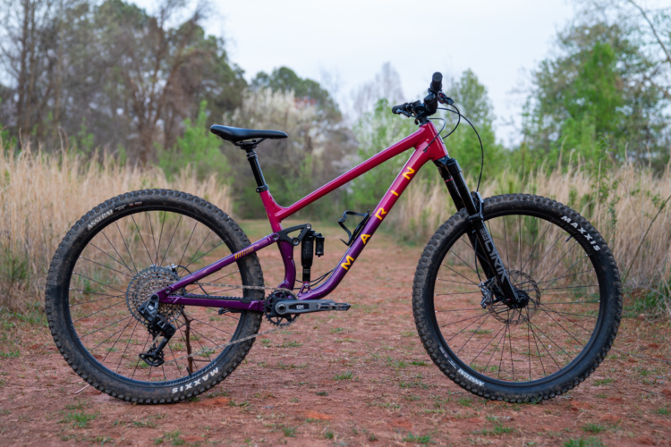 A purple mountain bike parked on a dirt path surrounded by grass and trees. The bike features thick tires, a suspension system, and is positioned to show its frame and components clearly.