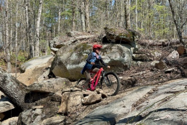 A mountain biker navigating a rocky trail in a wooded area, wearing a helmet and protective gear, with large boulders and trees in the background. South Rockdale Community Park mountain bike trail.