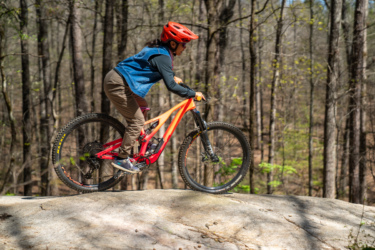 A person riding a mountain bike on a rocky terrain in a wooded area, wearing a red helmet and blue vest, with a focus on maintaining balance while navigating the obstacle. South Rockdale Community Park mountain bike trail.
