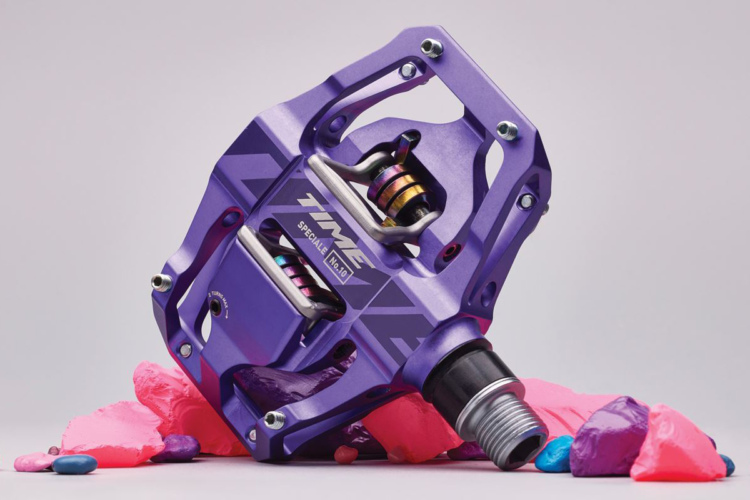 A close-up of a purple bicycle pedal set against a light gray background, with colorful modeling clay in pink, blue, and purple scattered around it. The pedal features a design with visible mechanical components and branding details.