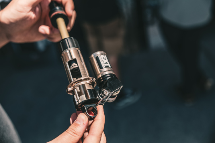 A close-up image of a person's hand holding a bicycle suspension component, showcasing a metal shock absorber with various mechanical features and a black attachment point. The background is slightly blurred, indicating that the setting is likely an event or workshop related to cycling or bicycle maintenance.