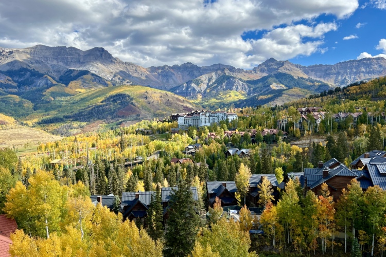 A scenic view of a mountain landscape featuring vibrant autumn foliage, showcasing a mix of green and golden trees. In the foreground, several rustic buildings with dark roofs are nestled among the trees, while in the background, majestic mountains rise under a partly cloudy sky. A resort building is visible on the hillside, surrounded by colorful fall foliage.
