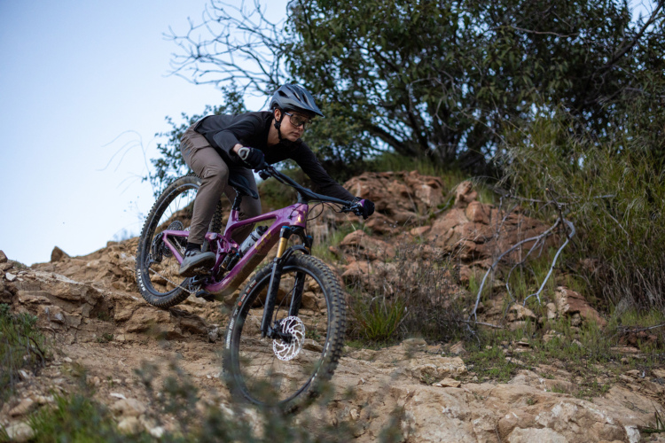 A mountain biker in a black long-sleeve shirt and helmet is skillfully navigating rocky terrain on a purple mountain bike, showcasing an action shot in a natural outdoor setting. The background includes rocky outcrops and sparse vegetation, with a clear blue sky overhead.