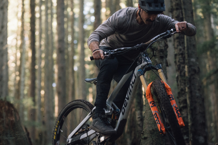 A mountain biker performs a trick on their bike amidst tall trees in a forest. The rider is wearing a helmet and is focused on the trail, with the bike's front wheel lifted off the ground. The scene captures the natural beauty of the woods, highlighting the action and energy of mountain biking.