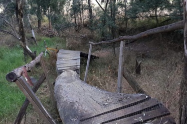 A wooden pathway winding through a forested area, with grass and trees in the background. A bicycle is leaning against the edge of the path. The structure appears rustic, made from logs and planks, and is surrounded by natural vegetation. Benoni Country Club MTB mountain bike trail.