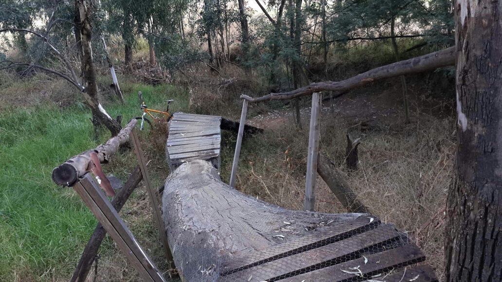 A wooden pathway winding through a forested area, with grass and trees in the background. A bicycle is leaning against the edge of the path. The structure appears rustic, made from logs and planks, and is surrounded by natural vegetation. Benoni Country Club MTB mountain bike trail.