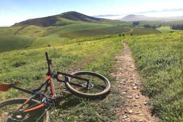 A scenic view of a green landscape with rolling hills in the background. In the foreground, an off-road bicycle is resting on a gravel path that winds through lush grass. The sky is clear, and the horizon features distant mountains shrouded in mist, suggesting an early morning or late afternoon setting. Contermanskloof mountain bike trail.