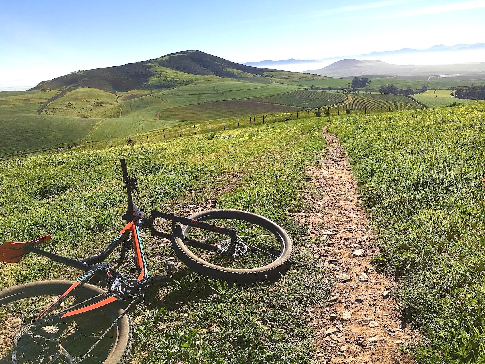 A scenic view of a green landscape with rolling hills in the background. In the foreground, an off-road bicycle is resting on a gravel path that winds through lush grass. The sky is clear, and the horizon features distant mountains shrouded in mist, suggesting an early morning or late afternoon setting. Contermanskloof mountain bike trail.