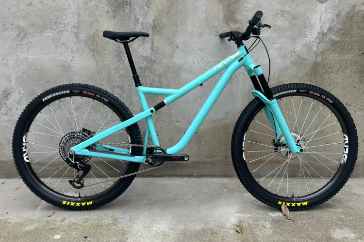 A turquoise mountain bike positioned against a textured gray wall. The bike features wide tires and a sleek frame with a prominent suspension fork, showcasing its design for off-road use. The background is a simple concrete surface.