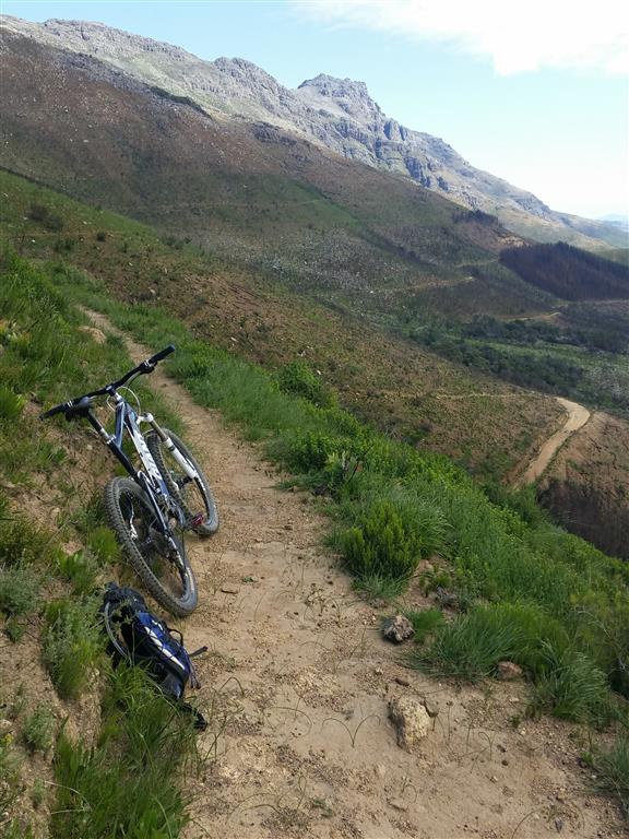 A mountain bike resting on a dirt trail surrounded by lush green grass and hills, with rocky mountains in the background and a winding path disappearing into the distance. Firehut Trail mountain bike trail.