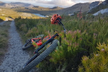 A mountain bike with a bright orange helmet rests on the ground beside a gravel path, surrounded by green foliage and blooming wildflowers. In the background, rolling hills and a lake create a scenic outdoor landscape under a partly cloudy sky. Hilcrest mountain bike trail.