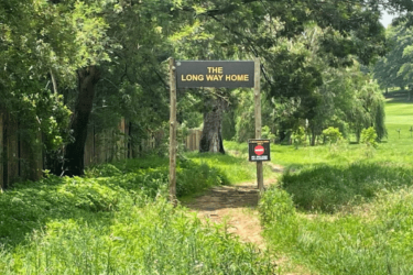 A grassy path lined with trees, featuring a sign that reads "THE LONG WAY HOME," along with a smaller sign below it. The area appears lush and green, evoking a sense of tranquility and nature. Huddle Park Golf & Recreation mountain bike trail.
