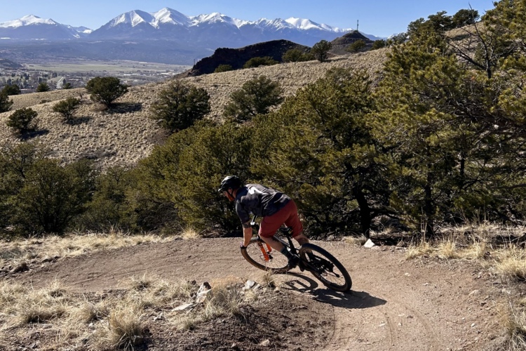 A mountain biker riding along a dirt trail with a smooth curve, surrounded by grass and sparse vegetation, against a backdrop of snow-capped mountains and a clear blue sky.