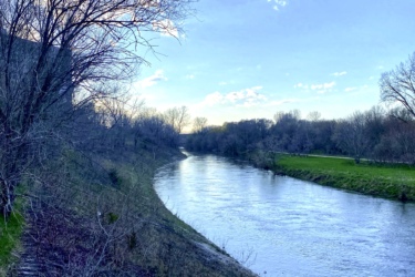A serene landscape view of a winding river surrounded by bare trees and lush greenery under a blue sky with scattered clouds. The riverbank features a stone path leading along the water's edge, reflecting the tranquility of the natural setting. MeadowLilly mountain bike trail.