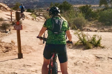 A person on a mountain bike, wearing a helmet and a green patterned shirt, is riding along a rocky trail with a distant view of cliffs and valleys. Another cyclist is seen in the background, and a sign indicating the suggested riding direction is positioned on the left. The sky is partly cloudy, creating a bright and dynamic outdoor scene. Getaway mountain bike trail.