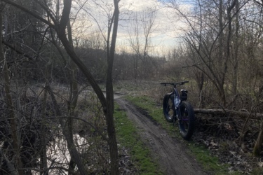 A fat bike parked on a dirt trail surrounded by sparse trees and overgrown vegetation. The setting is a natural outdoor area with a small body of water reflecting the sky. The sky is partly cloudy, suggesting a tranquil atmosphere. Bonneyville Mill mountain bike trail.
