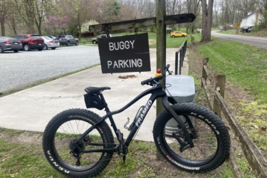 A fat-tire bike parked near a sign that reads "Buggy Parking." The bike is resting next to a trash can, with a gravel parking area and cars visible in the background. Trees and a road are seen in the distance, creating a natural setting. Bonneyville Mill mountain bike trail.