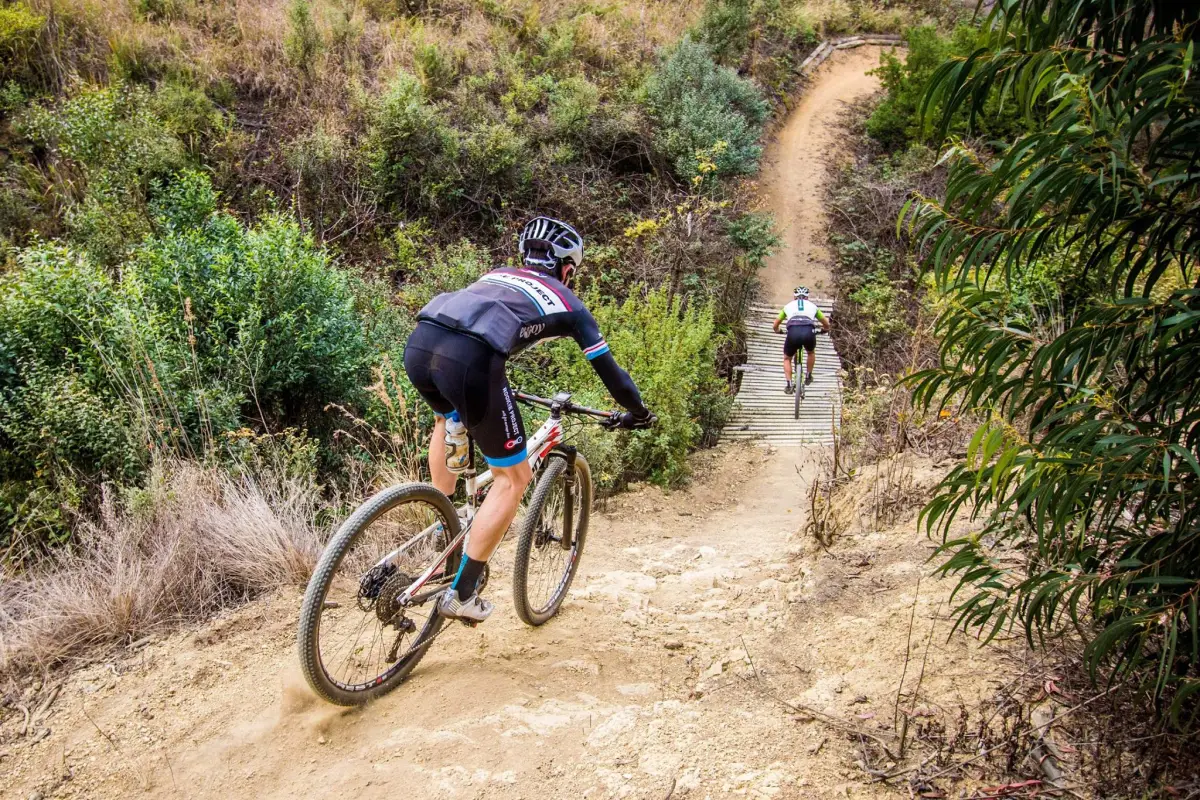 Two mountain bikers navigate a dirt trail surrounded by greenery. The cyclist in the foreground rides downhill on a rocky path, while the second cyclist ascends a wooden ramp in the background. The landscape features a mix of bushes and dry grass, with a winding trail leading off into the distance. Karkloof to Howick and back mountain bike trail.