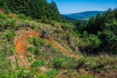 A mountain biker navigating a dirt trail on a lush hillside, surrounded by greenery and distant mountains under a clear blue sky. Karkloof 30km mountain bike trail.