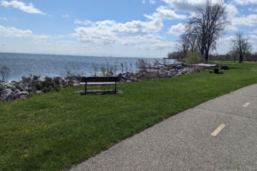 A scenic view of a waterfront path lined with grass, featuring a wooden bench facing the water. To the left, rocks outline the shore, and in the background, a calm lake reflects a bright blue sky with fluffy white clouds. Sparse trees are visible in the distance. Lake Erie XC Trails mountain bike trail.