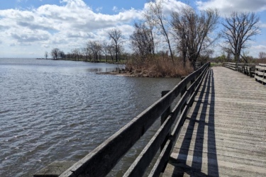A wooden boardwalk stretches along the water's edge, curving toward a tree-lined shore under a partly cloudy sky. The calm water reflects the clouds above, while trees on the bank show signs of early spring with sparse leaves. Shadows from the railing create a pattern on the walkway. Lake Erie XC Trails mountain bike trail.