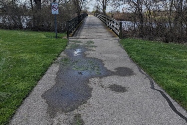 A paved pathway leading to a wooden bridge over a waterway, surrounded by greenery and bare trees. Puddles are visible on the path, and a no-trespassing sign is posted nearby. The sky is partly cloudy, indicating a clear day. Lake Erie XC Trails mountain bike trail.