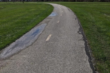 A winding pathway through a green park, with patches of water visible on the asphalt. The sky is partly cloudy, and trees line both sides of the path, showcasing bare branches and fresh foliage. Lake Erie XC Trails mountain bike trail.