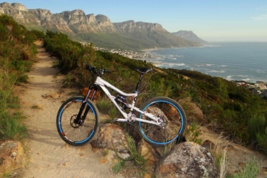 A mountain bike resting on a rocky path with lush greenery on either side, overlooking a scenic coastal landscape with cliffs and the ocean in the background. Lions Head mountain bike trail.