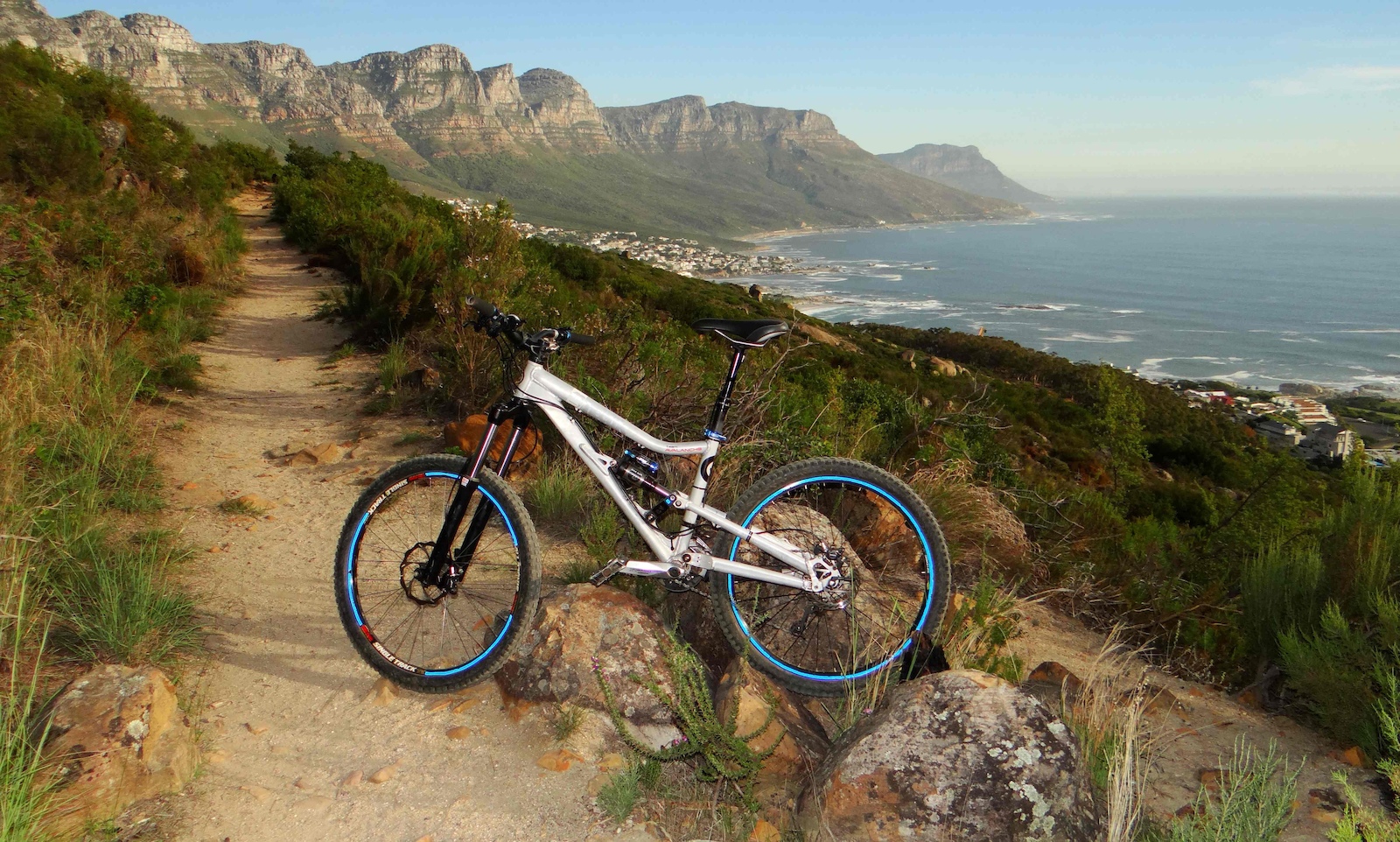 A mountain bike resting on a rocky path with lush greenery on either side, overlooking a scenic coastal landscape with cliffs and the ocean in the background. Lions Head mountain bike trail.