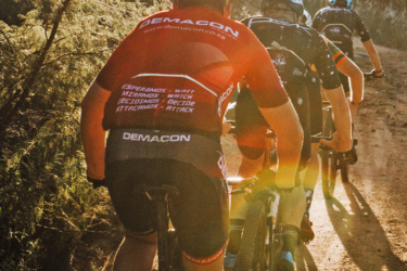 A group of cyclists riding on a dirt path during sunset. The front cyclist wears a red jersey with the logo "DEMACON," while others follow closely behind, dressed in various cycling attire. Sunlight filters through the trees, creating a warm and dynamic atmosphere. Northern Farms mountain bike trail.