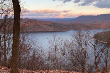 A scenic view of a lake surrounded by trees during autumn, with fallen leaves on the ground. The sky is partly cloudy, displaying hues of orange and pink from a sunset in the background, casting a tranquil atmosphere over the landscape. Allegrippis Trails mountain bike trail.