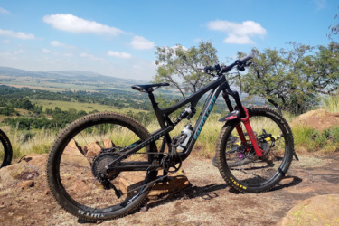 A mountain bike resting on rocky terrain, with a scenic view of rolling hills and a blue sky dotted with clouds in the background. The bike features a black frame with colorful accents and is equipped for off-road riding, showcasing its rugged tires and suspension. Thaba Trails mountain bike trail.