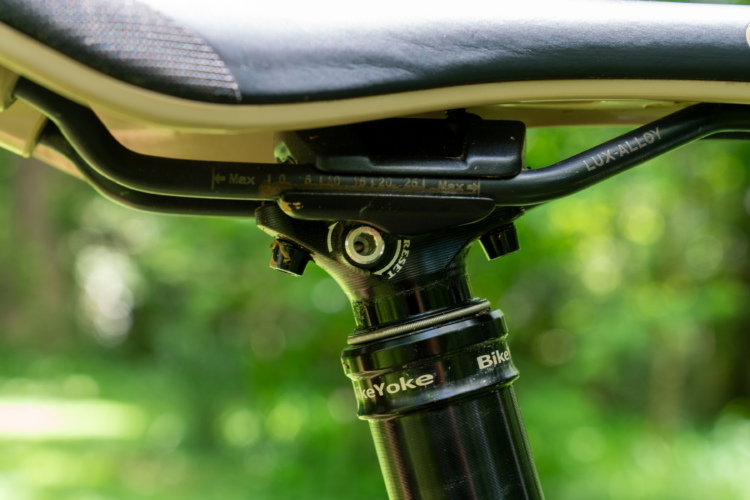 Close-up view of a mountain bike seat post and saddle, showcasing the mounting mechanism and branding. The background is a blurred green natural setting, suggesting an outdoor environment.
