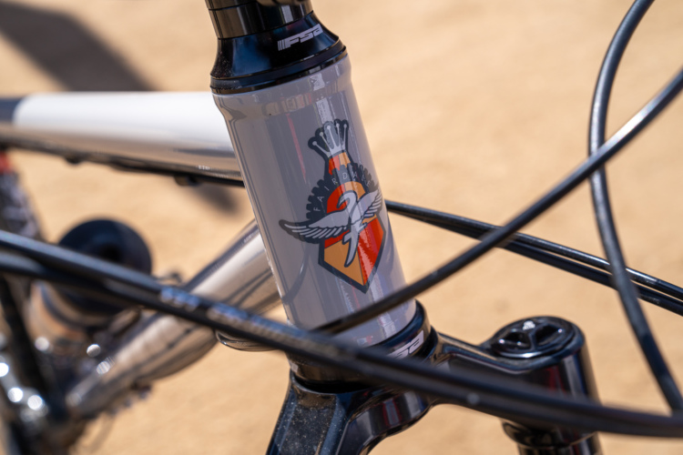 Close-up image of a bicycle's head tube featuring a logo with a flying bird and a crown, set against a light sandy background. The bike frame is metallic with intricate detailing and black cables visible in the foreground.