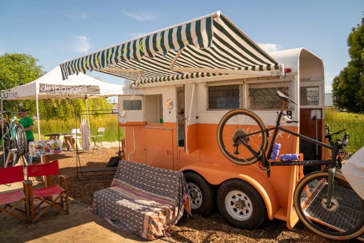A colorful food trailer with a striped awning, parked in a outdoor setting. In front, there is a bicycle with a water bottle attached, and two wooden chairs. A vendor in a green shirt is working at a nearby table with bike accessories, under a tent labeled "dirtbaggies." The background features tall grass and trees under a clear blue sky.