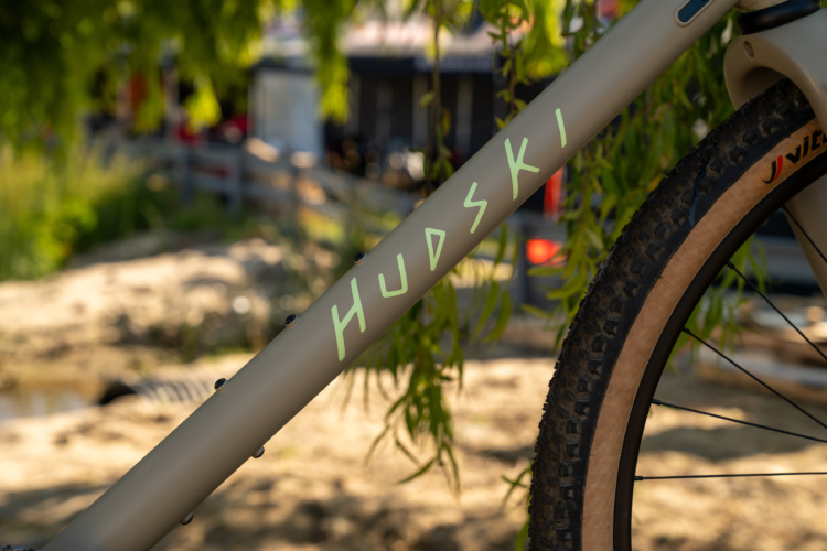 Close-up of a bicycle frame featuring the brand name "Hudski" in a light green font. The bike is positioned in a natural outdoor setting, with greenery in the background and sandy ground visible. The image captures details of the bike's design, including the textured tire and part of the braking system.