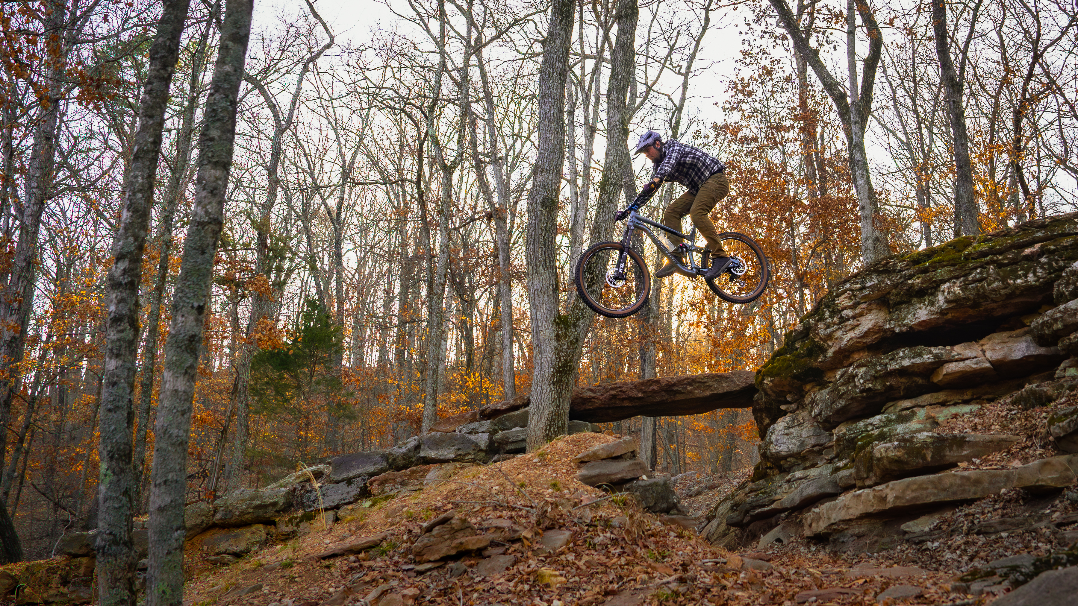 A mountain biker performs a jump over a rocky outcrop in a forest during autumn, surrounded by trees with colorful leaves.