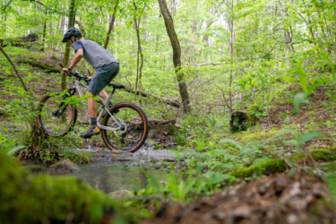 A person riding a mountain bike over a shallow stream in a lush, green forest during daytime. The cyclist is wearing a helmet and appropriate biking attire, navigating through a natural setting filled with trees and foliage. Lake Ouachita Vista Trail (LOViT) mountain bike trail.