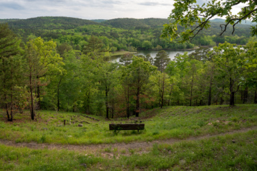 A tranquil view of a green landscape featuring a park bench overlooking a serene lake, surrounded by lush trees and rolling hills under a cloudy sky. Lake Ouachita Vista Trail (LOViT) mountain bike trail.