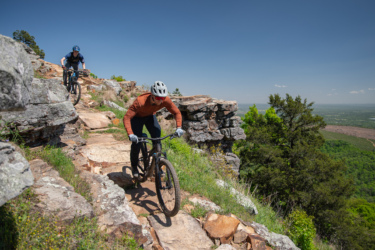 Two mountain bikers navigate a rocky trail on a hillside under a clear blue sky. One rider is in the foreground, wearing an orange long-sleeve shirt and a white helmet, while the other is visible in the background, dressed in a blue shirt and helmet. The landscape features greenery and rocky outcrops, with a scenic view stretching into the distance. Mount Nebo State Park mountain bike trail.