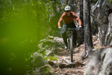 A person riding a mountain bike on a narrow trail surrounded by trees and rocky terrain. The rider is wearing a helmet and sunglasses, dressed in an orange long-sleeved shirt and black pants, navigating the uneven path amidst a lush green environment. Mount Nebo State Park mountain bike trail.