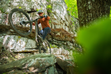 A mountain biker performs a stunt on a rocky terrain, navigating a steep incline as the front wheel of the bike is lifted off the ground. Lush greenery surrounds the area, with large rock formations and trees in the background. The rider is wearing protective gear, including a helmet and gloves, and is focused on the challenging path ahead. Mount Nebo State Park mountain bike trail.