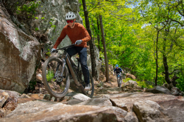 A mountain biker navigating a rocky trail in a lush green forest. The rider in the foreground is lifting the front wheel of their bike over large stones, while another cyclist follows behind on a winding path surrounded by trees. Bright sunlight filters through the leaves, creating a vibrant outdoor atmosphere. Mount Nebo State Park mountain bike trail.