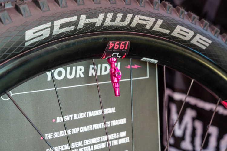 Close-up of a mountain bike wheel featuring a Schwalbe tire, a black rim, and a bright pink valve. In the background, visible text highlights features such as a silicone top cover that protects a tracker, emphasizing performance and ease of use for riders.