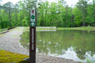 Signpost marking Sanderson Pass, located near a calm pond surrounded by lush greenery and trees. The sign indicates that the trail is the "easiest" and features a directional arrow pointing straight ahead. The path is gravely with some moss visible on the ground. Northwoods Trail System mountain bike trail.